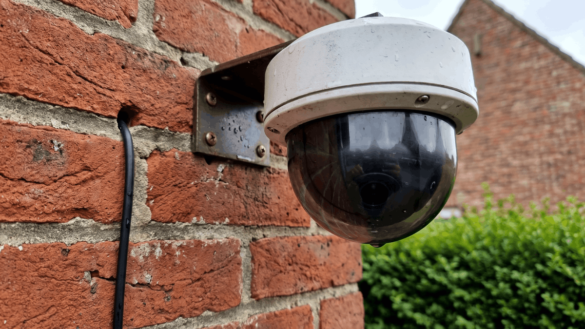 Een close-up toont een witte koepelcamera op een bakstenen gevel waarbij de donkere getinte koepel verbergt in welke richting de lens kijkt en de kabel strak langs de voeg naar beneden loopt