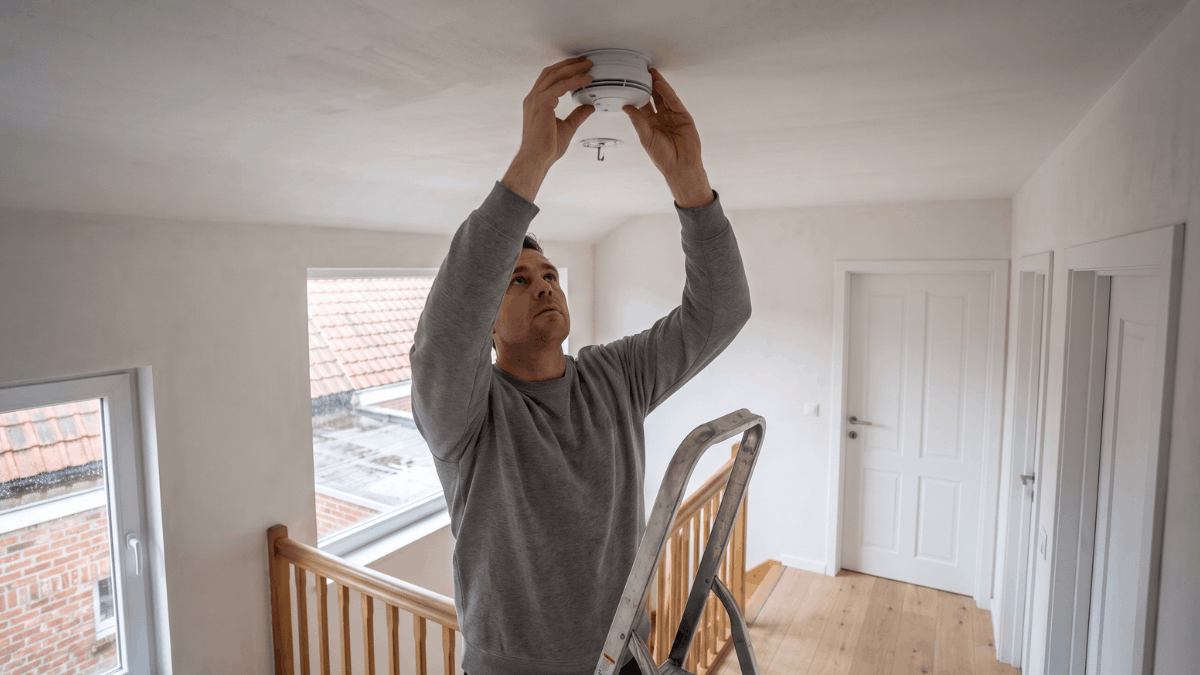 Een man op een trapladder duwt een ronde witte rookmelder tegen het plafond midden op de overloop van een rijwoning.