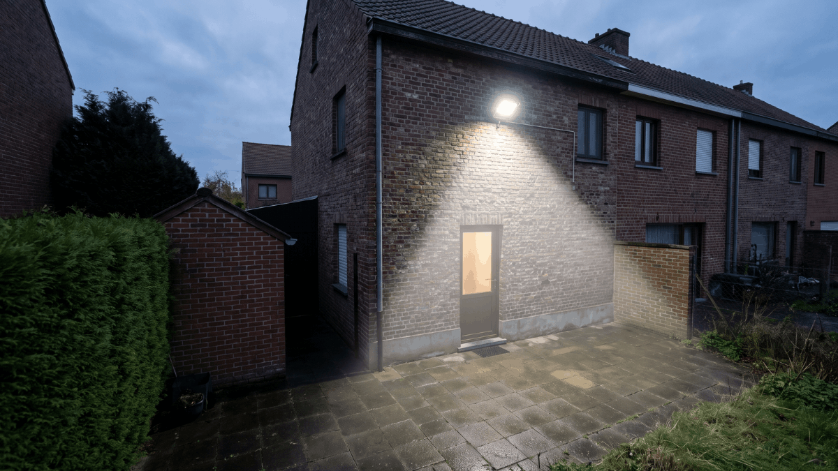 Een breedstraler op de achtergevel verlicht het terras terwijl de hoek naast de tuinberging in het donker blijft.