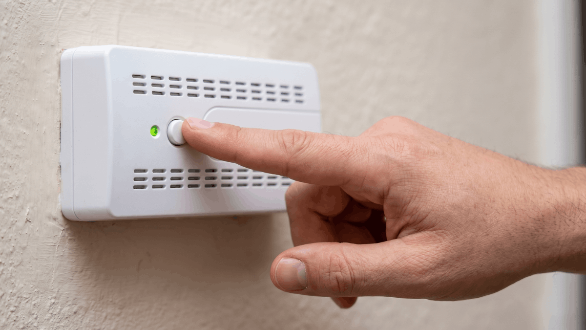 Een wijsvinger drukt de ronde testknop in op een witte gasmelder aan de muur terwijl het groene indicatielampje ernaast brandt.
