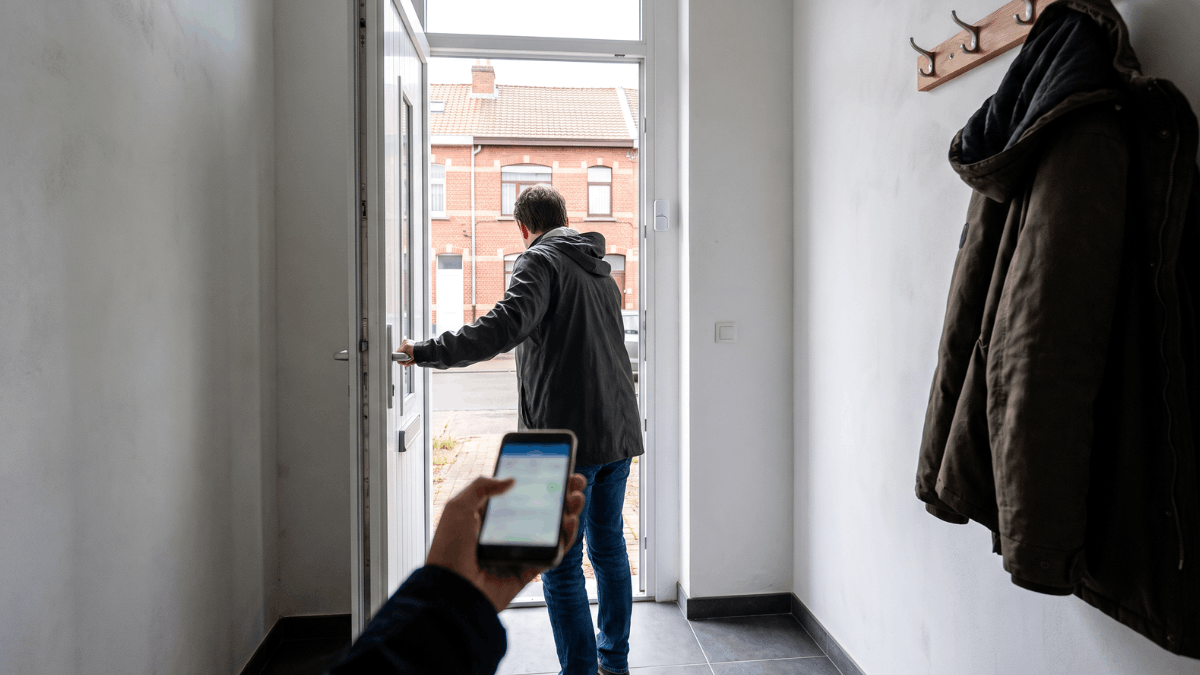 Een bewoner stapt de hal van een Belgische rijwoning binnen met een smartphone in de hand terwijl op het deurkozijn een klein wit magneetcontact zichtbaar is.