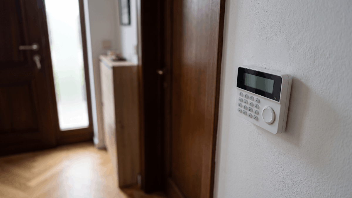 Een compact wit alarmbedienpaneel met cijfertoetsen en badgelezer hangt op ooghoogte naast een donkere houten voordeur in een Belgische hal met lichte parketvloer.